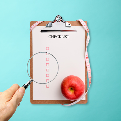 Close-up of looking through a checklist with a magnifying glass, on light blue background.
Red apple on the checklist.
Dieting concept