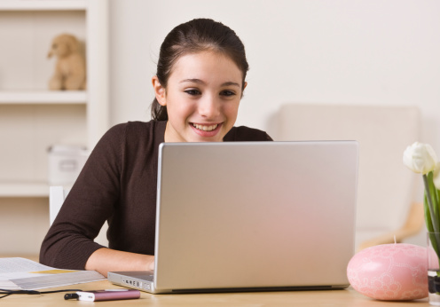 A young girl is working on a laptop, smiling, and looking away from the camera.  Horizontally framed shot.  
