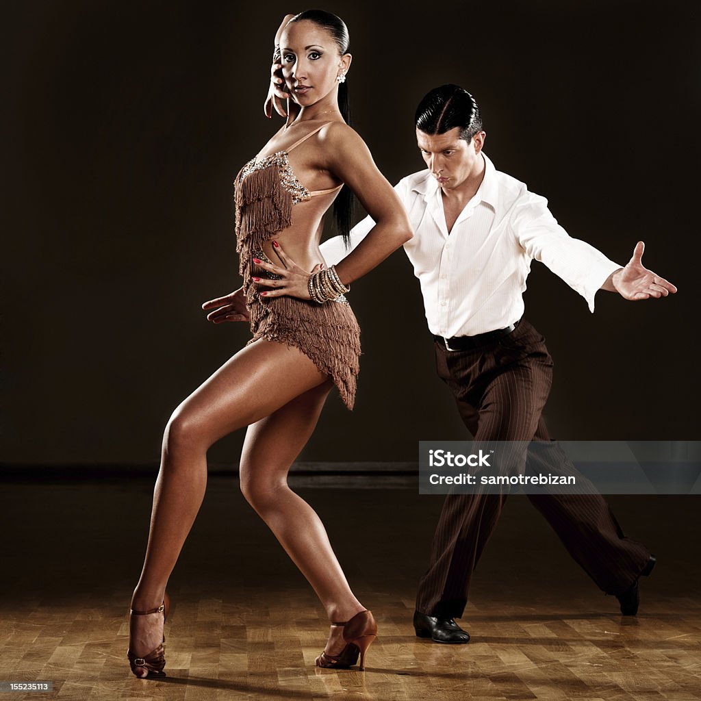 Professional dancing couple performing in ballroom Latino dance couple dancing in dark ballroom with wooden floors. He is wearing white shirt and black trousers and she light brown dress. Both standing in the dance pose, he is behind her and looking in the floor. Dancing Stock Photo