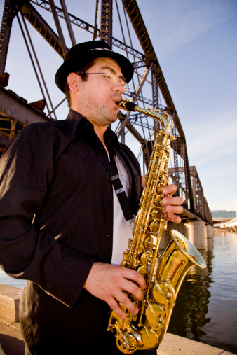 A saxophonist plays outdoors at sunset against a grungy industrial skyline.