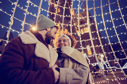 Happy couple in love holding hands while communicating during Christmas night in the city.