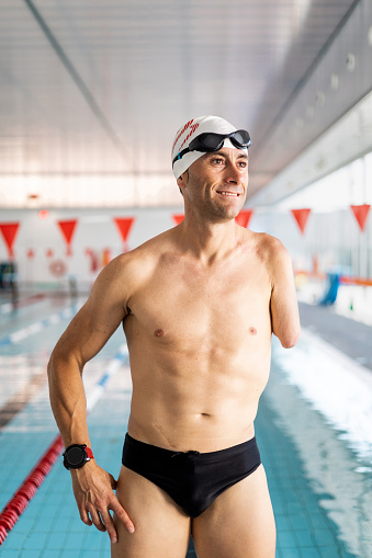 Portrait of an adult swimmer with an amputated arm is posing outside an indoor pool while looking to the side happily. Concept of disabled swimmers, athletes with an amputated arm. Motivation and determination.