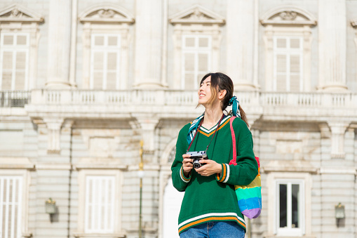 Exuberant Oriental Traveler Capturing Memories with Camera in Historic City, Carrying Gay Pride Bag