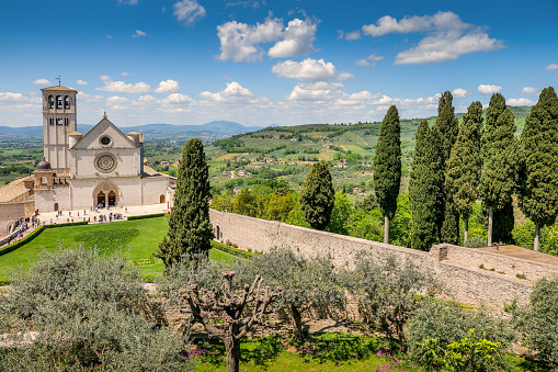 A beautiful and peaceful landscape with the Basilica di San Francesco (Basilica of St Francis) in the medieval town of Assisi, in Umbria, surrounded by olive trees and Italian cypresses. Built in the Italian Gothic style starting from 1228 and completed in 1253, the Basilica, which preserves the mortal remains of the Saint of the Poor from 1230, is composed of the Basilica Inferiore (Lower Basilica) and the Basilica Superiore (Upper Basilica), perfectly integrated. Over the centuries Assisi and the spirituality of its sacred places have become a symbol of peace, a point of reference for tolerance and solidarity between peoples and between the different confessions of the world. The Umbria region, considered the green lung of Italy for its wooded mountains, is characterized by a perfect integration between nature and the presence of man, in a context of environmental sustainability and healthy life. In addition to its immense artistic and historical heritage, Umbria is famous for its food and wine production and for the high quality of the olive oil produced in these lands. Since 2000 the Basilica and other Franciscan sites of Assisi have been declared a World Heritage Site by UNESCO. Super wide angle image in high definition quality.