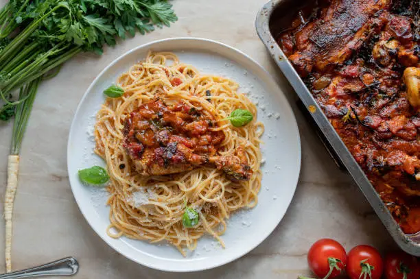 Delicious italian braised chicken. Cooked with tomatoes, vegetables and herbs. Served with spaghetti and grated parmesan cheese on a plate with roasting pan on the side. Top view