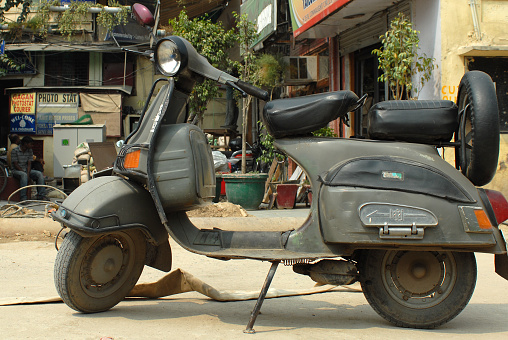 Urban downtown life and a dusty moped parked in the foreground on a sunny afternoon in the streets of New Delhi, India