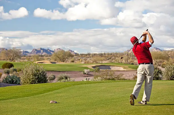A golfer on the tee on a beautiful desert golf course. Mesa, Arizona. Horizontal  colour image. Caucasian male. 30s. Back view of professional left-handed golfer on the tee box on a pristine golf hole in the Sonoran desert. Golfer is making a perfect balanced and athletic swing. Themes of this image include: golfer, male, white, Arizona, united states, balance, fitness, health, senior, men, turf, model, red, pose, winter, snowbird, back view, and unrecognizable people. One man is in the image. 