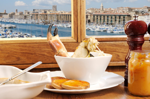 Traditional serving of Marseille bouillabaisse (fish stew) , served with rouille on toast. Through the old-fashioned wood-framed window is a view of the old harbour, where there is a daily fish market, Marseille fish stew consists of the catch of the day (fish and shellfish). Heads, tails and trimmings are covered in water, with chopped onion, garlic, leek, salt and pepper, and boiled for twenty minutes, then strained to make the bouillon (fish stock). Chopped fennel, onion, skinned tomatoes and garlic are then turned in oil and simmered till tender, when pieces of fish are added along with strands of saffron. Marseille bouillabaisse is traditionally served with rouille, a spicy paste spread on toasted baguette. The soup is sometimes served as a starter, with the large fish pieces as the main course.
