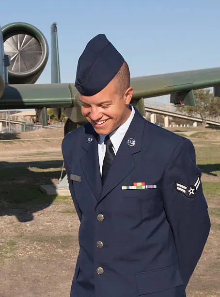 Airman in dress blue uniform smiles casually in front of airplane display. Lackland Air Force Base graduation weekend. Airman has ribbons and 2 strips. Name has been removed from name tag. Happiness and pride show in this casual portrait.
