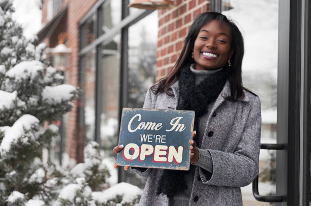 Smiling African American woman in front of storefront holding an open sign on a winter day. Shot in downtown DeWitt, Michigan.