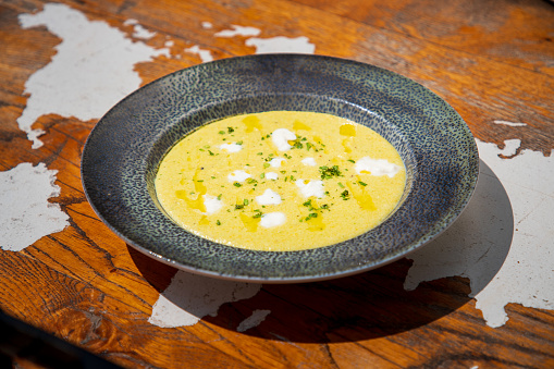 Yellow potage served with cream scoops and chopped parsley on top in plate, wooden table in fine dining