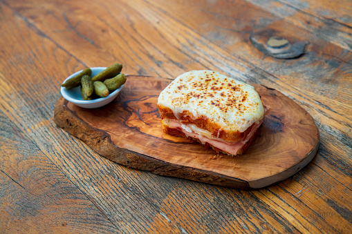Juicy ham sandwich with melting cheese on top, served on wooden board with pickles in bowl on restaurant table, fine dining snack