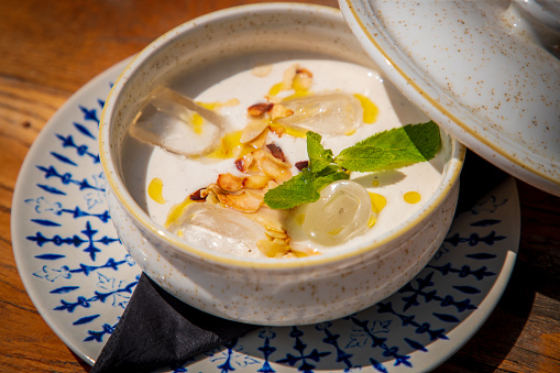 White colored potage served in bowl with open lid, cooled with ice and garnished with mint leaves and almond flakes, fine dining close-up