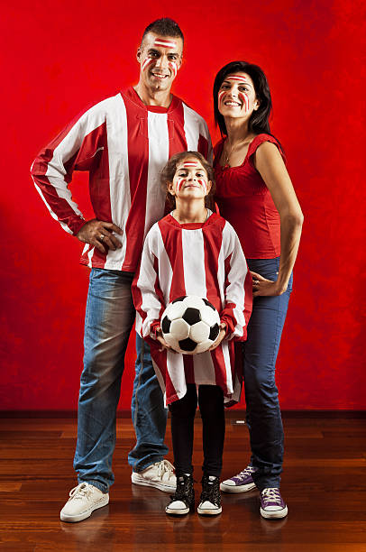 full length portrait of a family of soccer fans wearing their team color posing against a red wall.