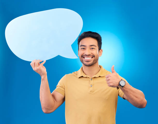 istock Young man, speech bubble and studio portrait for thumbs up, opinion and social network by blue background. Indian guy, student or model for yes icon, mock up space and cardboard sign for promotion 1552817620