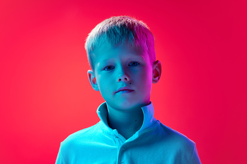 Portrait of little boy, child with short blonde hair posing in white shirt against pink studio background in neon light. Concept of childhood, lifestyle, emotions, education, fashion, care, ad