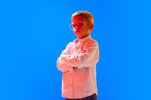 Portrait of smart, serious boy, child, pupil standing in white shirt and glasses against blue studio background in neon light. Concept of childhood, lifestyle, emotions, education, fashion, care, ad