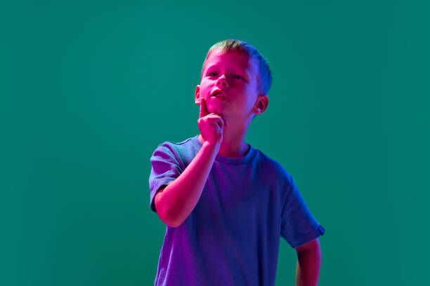 Portrait of little smart boy, child standing with thoughtful face against cyan studio background in neon light Portrait of little smart boy, child standing with thoughtful face against cyan studio background in neon light. Concept of childhood, lifestyle, emotions, education, fashion, care, ad neon portrait boy stock pictures, royalty-free photos & images