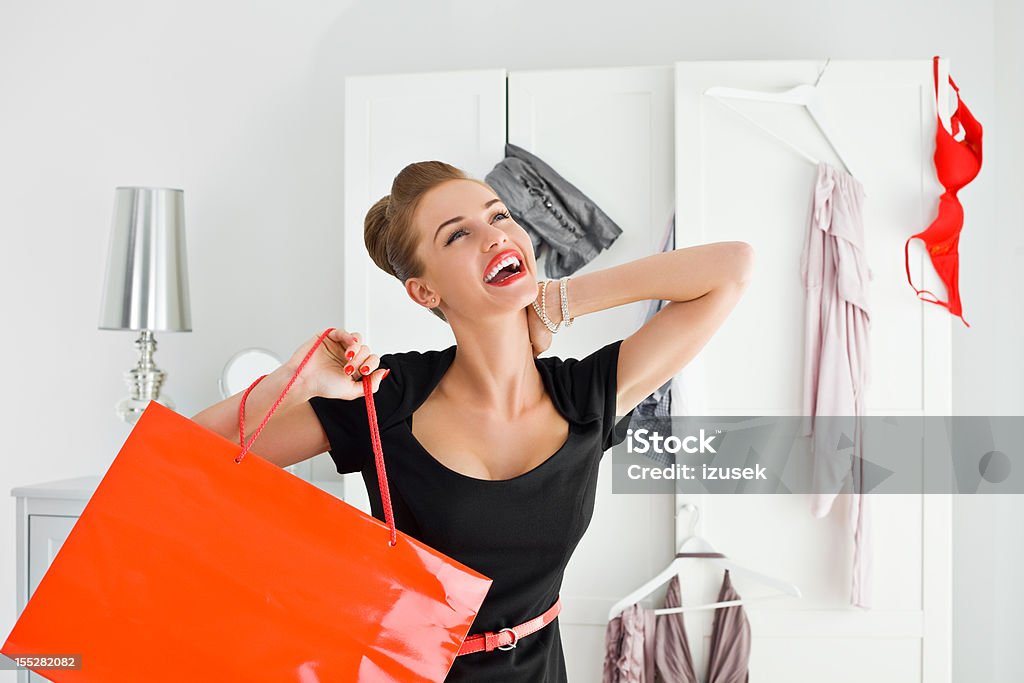 Beautiful shopaholic Portrait of happy young adult woman holding red shopping bag in her hand with messy wardrobe in the background.  Happiness Stock Photo
