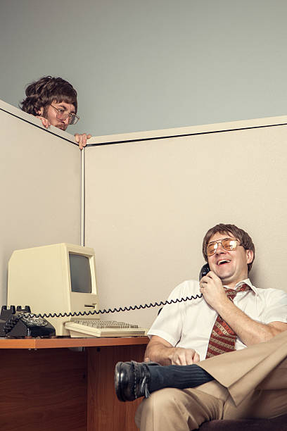 A manager spies over an office cubicle on his co-worker, looking very angry.  The other business man is on a phone call laughs, presumable slacking off, OR talking bad about his manager.  1980's styling, complete with vintage computer.  Vertical with some copy space.
