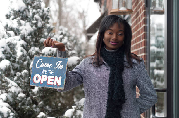Smiling African American woman in front of storefront holding an open sign on a winter day. Shot in downtown DeWitt, Michigan.