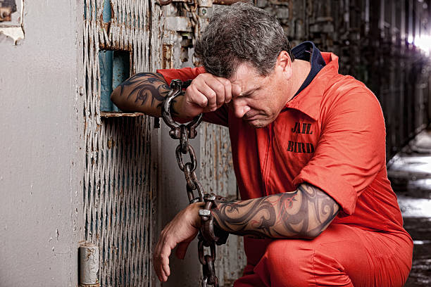 A tattooed death row prisoner, in shackles, kneels and prays as his time is drawing near, hanging his head in remorse.