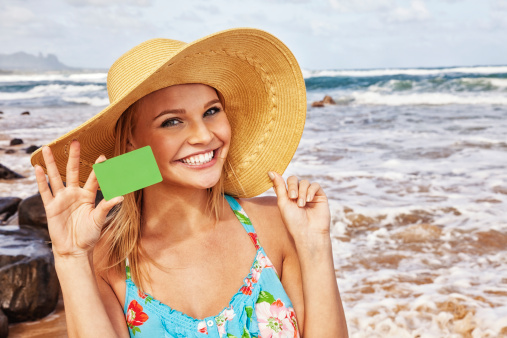 Photo of an attractive, happy young blonde woman on a Hawaiian beach, wearing a large floppy beach hat, holding a blank green credit card (or gift, reward, incentive, etc.).