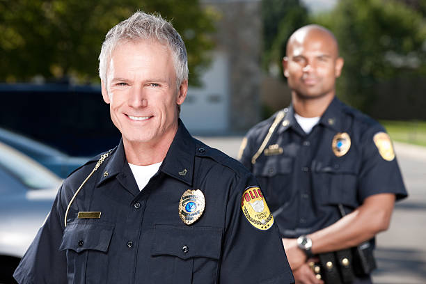 Mature Police Officer Portrait Head and shoulders portrait of a friendly law enforcement officer in his early 50s. His younger african american partner stands in the background behind him. police force smiling officer stock pictures, royalty-free photos & images