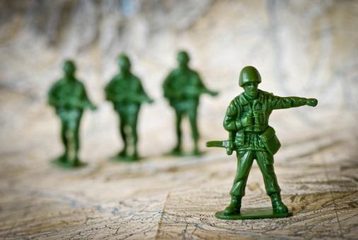 Square composition photography of concept of leadership, with little green soldier toy aiming the way forward, with arm raised, concept of teamwork in business. Macro picture shot, taken isolated on white background in studio. The dominant color is green khaki.