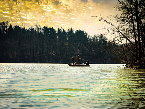 Two anonymous silhouetted men are fishing on a bass boat in this moody winter scene at sunrise or sunset.