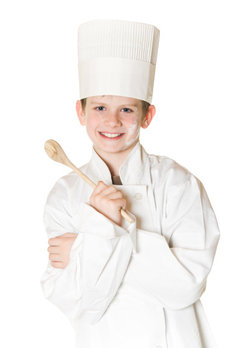 Happy smiling little kid with curly blond hair in white chef hat and jacket standing with ladle in hand in kitchen while cooking healthy food at home and looking at camera