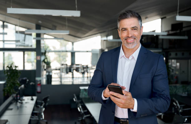 istock Happy middle aged business man ceo standing in office using smartphone. 1552851595