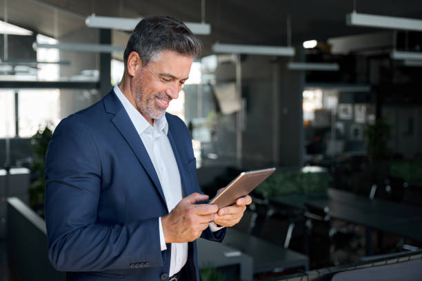 istock Happy mature business man wearing suit standing in office using digital tablet. 1552851611