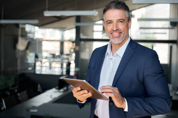istock Smiling professional business man wearing suit standing in office using tablet. 1552851622