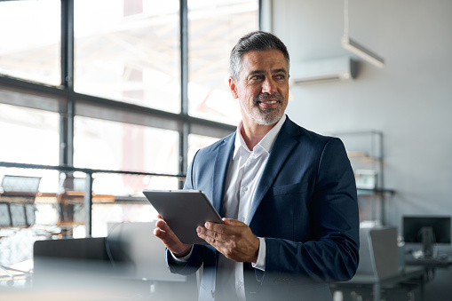 Happy middle aged business man ceo wearing suit standing in office using digital tablet. Smiling mature businessman professional executive manager looking away working on tech device. Authentic shot