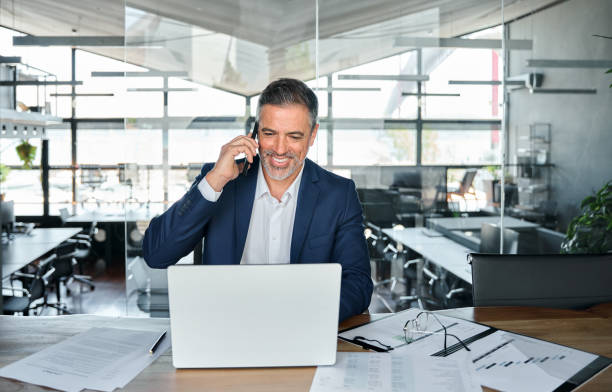 istock Smiling mature business man talking on cell phone using laptop in office. 1552851627