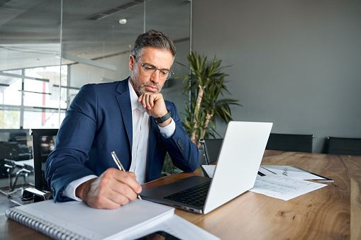 Busy serious middle aged business man executive sitting at desk working on laptop writing notes. Professional businessman ceo manager using computer for elearning or management in modern office.