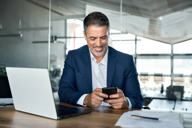 istock Happy mid aged business man wearing suit sitting in office using mobile phone. 1552851664