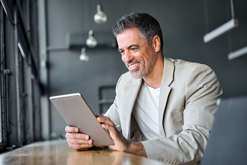 Happy mid aged business man wearing suit standing in modern office using digital tablet. Mature businessman professional manager holding tab working on financial data on fintech device.