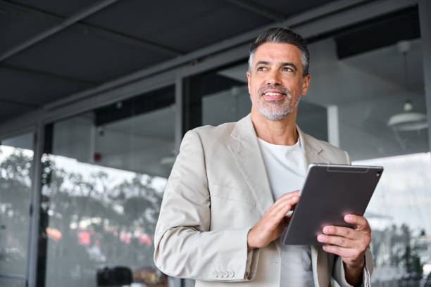 デジタルタブレットを持つオフィスの外に立つ笑顔の中年のビジネスマン。 - digital tablet people business outdoors ストックフォトと画像