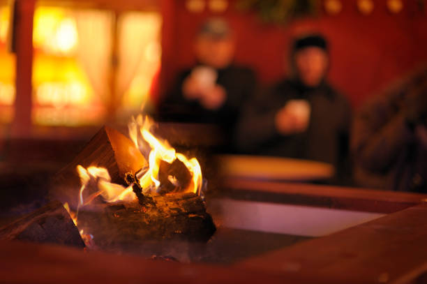 **Focus on fire with out of focus couple, in winter coats and hats, drinking from beakers at a table in the background** Cosy light from window of house but this couple have nowhere to go and are seeking warmth from a log fire outdoors