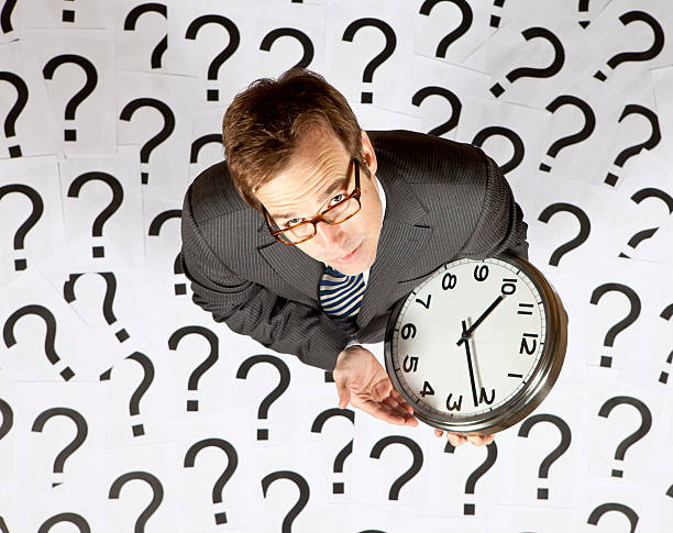 A business man holding a clock looks up at camera  Businessman holding clock surrounded by question marks calendar 2012 stock pictures, royalty-free photos & images