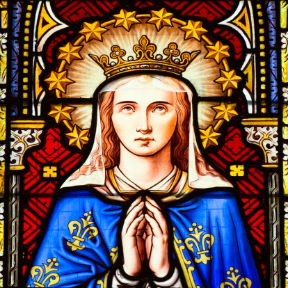 Beautiful stained glass window of cathedral Église de Nôtre-Dame du Sablon in Brussels. Woman with praying folded hands looking to sky and wearing a crown surrounded with halo and gloriole made of golden stars. Painting shows Blessed Virgin Mary: Queeen of Heaven. Her blue robe is decorated with lily flowers as sign og her purity and virginity. Gothic church is from 14th century. Restauration of cathedral began in 1870 by architect Auguste Schoy and work was followed by others til 1937.