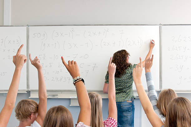 female high school teacher writing formula on whiteboard in classroom rear view on female high school teacher writing mathematical formula on whiteboard in classroom and students with raised hands teenage high school girl raising hand during class stock pictures, royalty-free photos & images