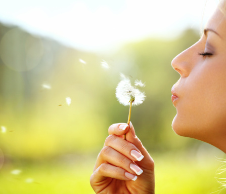 Closeup of woman blowing a dandelion against blurred green background. She's holding her eyes closed. It's hot summer day. Side view,low angle shot.