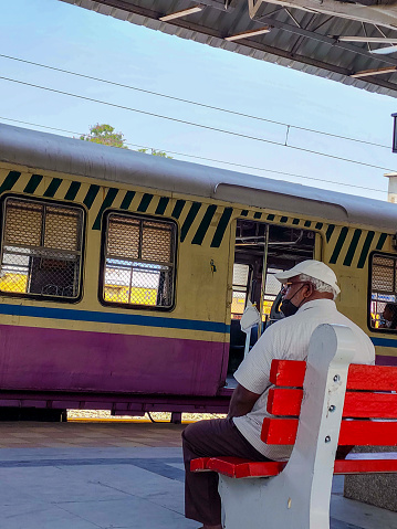 Hyderabad,India- October 22nd 2022; an Indian senior man wearing casual clothing, cap, and mask is seen sitting on a bench, patiently waiting for a train,with a local train arriving in the background