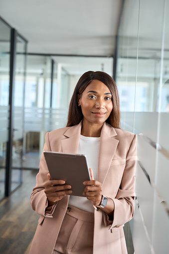 Young professional African American business woman company manager executive wearing suit holding fintech tab digital tablet computer standing in office at work, looking at camera, vertical portrait.