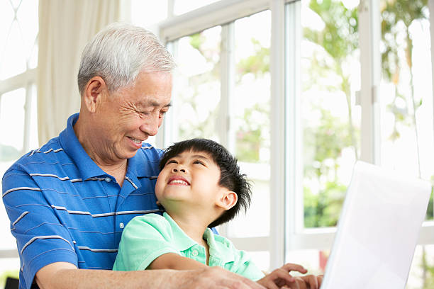 Chinese Grandfather And Grandson Sitting At Desk Using A Laptop Chinese Grandfather And Grandson Sitting At Desk Using Laptop At Home Smiling At Each Other china chinese ethnicity smiling grandparent stock pictures, royalty-free photos & images