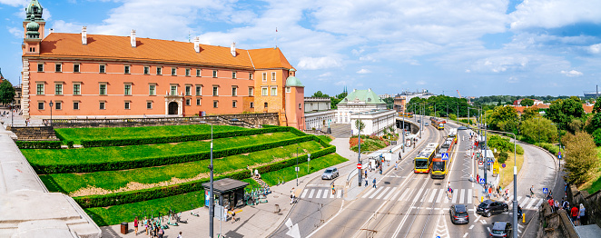 A view of Aleja Solidarności Street from Castle Square (Plac Zamkowy). The street is bustling with activity as people wait for public transport. Cars can be seen moving along the street. In the frame, a tram approaches a tram station, highlighting the public transportation system in the area. The photo offers a glimpse of the lively urban environment, showcasing the daily movements of people and vehicles on Aleja Solidarności Street.