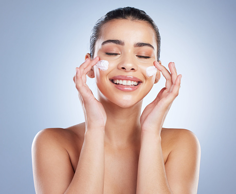 Skincare, hand and happy woman with face cream in studio for anti aging, wellness or hydration on grey background. Facial, smile and asian lady model with sunscreen, lotion or beauty mask application